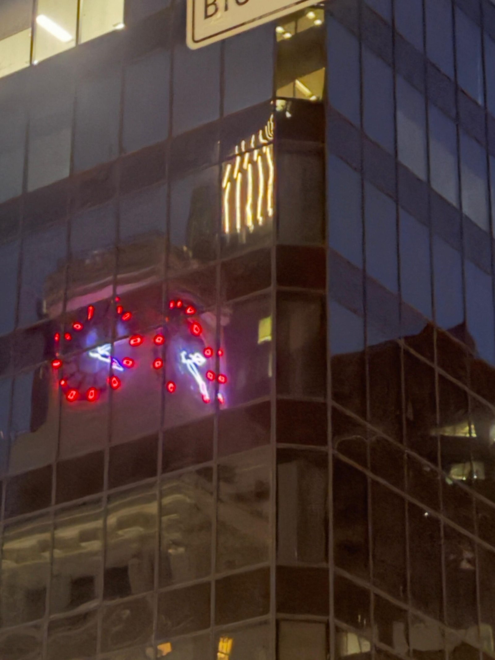 Red illuminated clocks reflected in the glass facade of a city building at dusk.