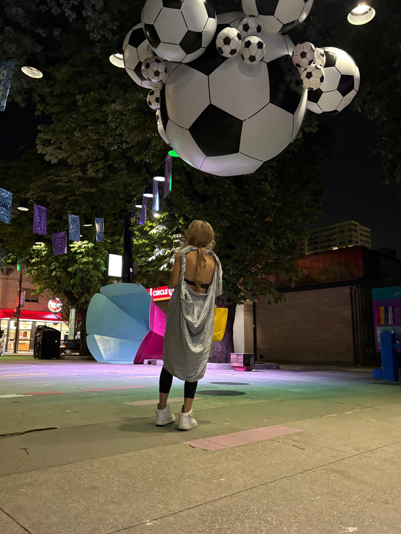 Woman standing beneath oversized suspended footballs in a city square at night
