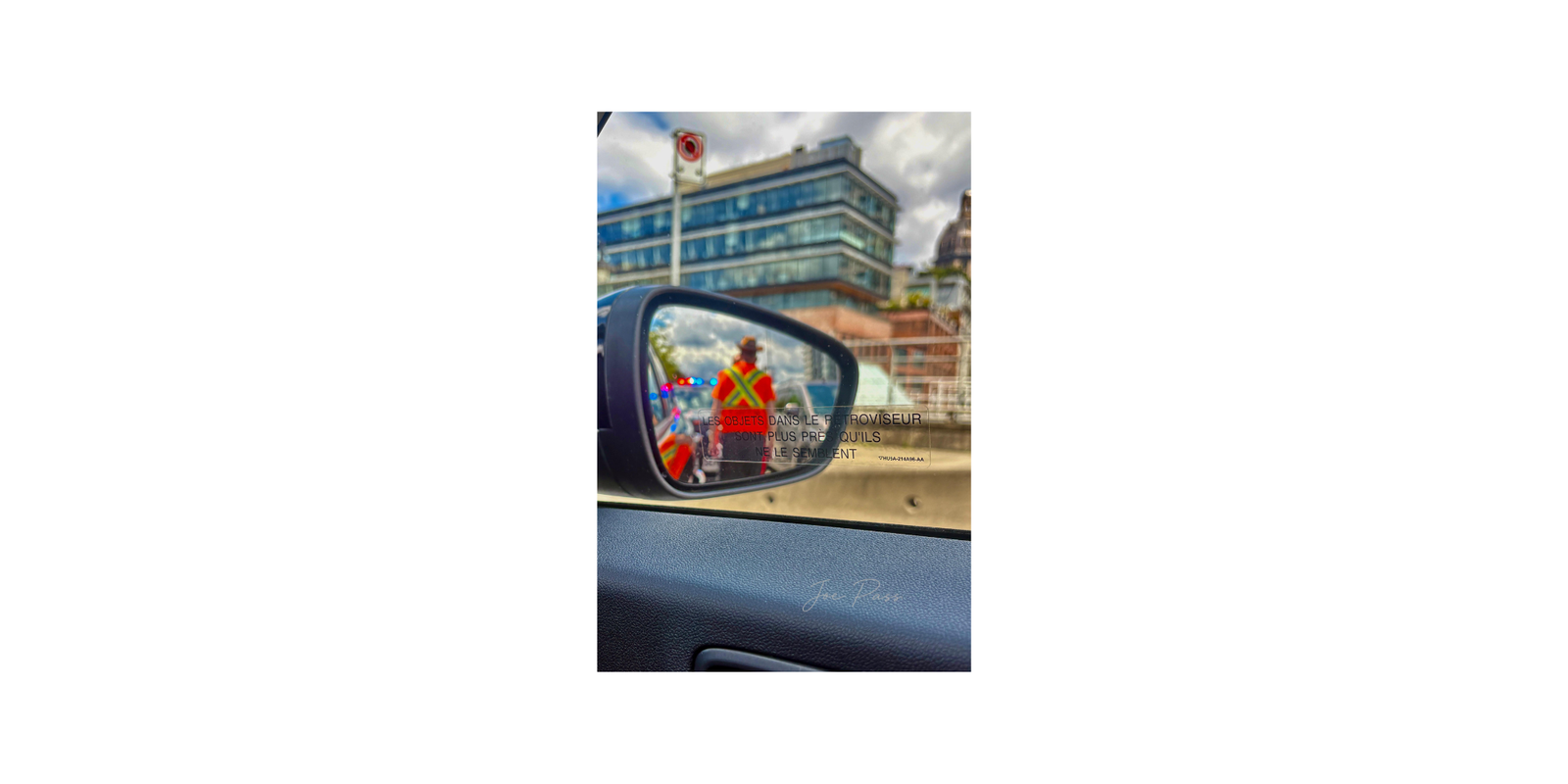 A road worker reflected in a car side mirror with city buildings and flashing lights in the background.